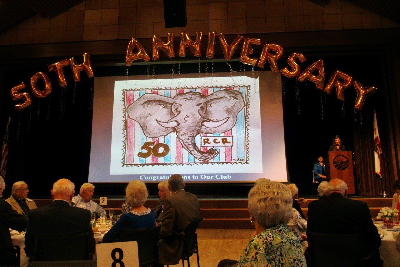 Anniversary event with audience seated before a stage showing a large screen and “50th Anniversary” banner