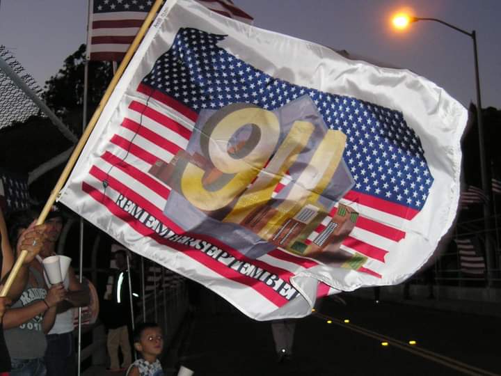 Large 9/11 flag being waved at a nighttime roadside vigil with people nearby