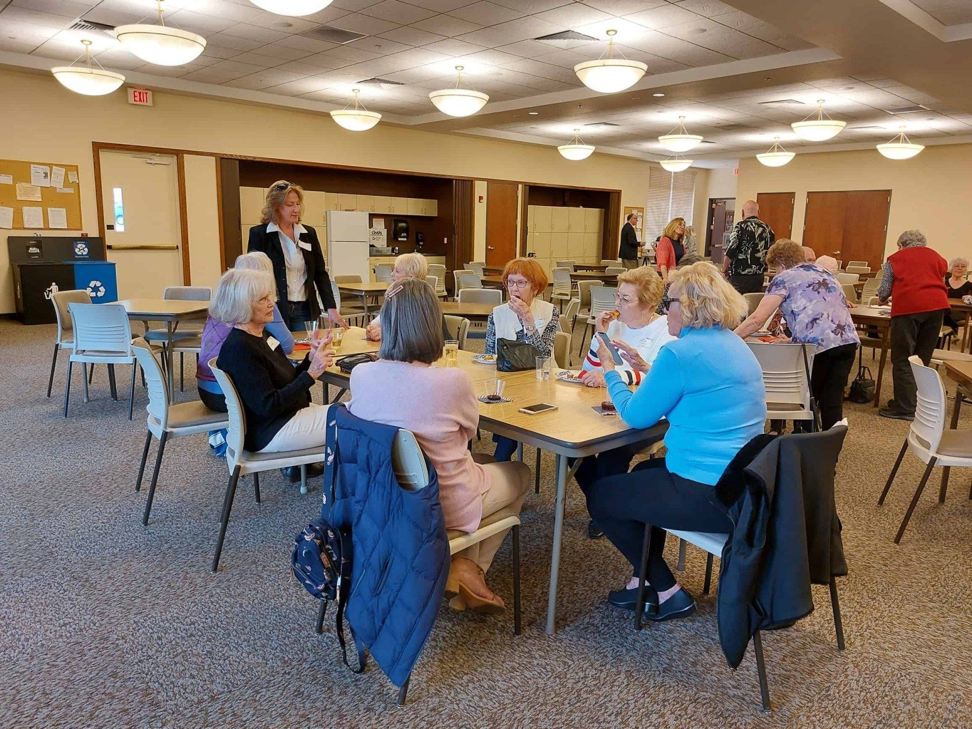People seated at round tables in a community room, chatting over drinks and snacks.