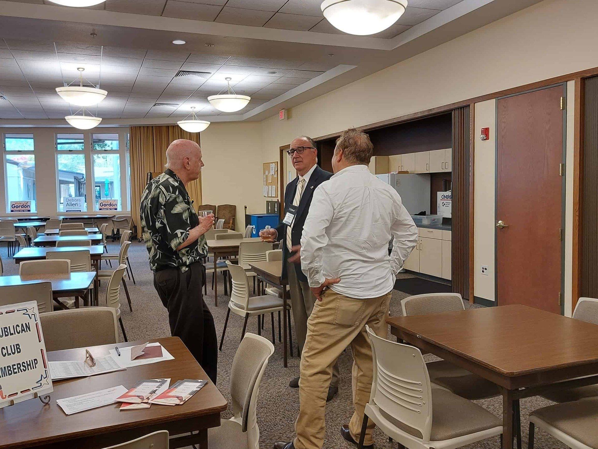 People talking in a meeting room with tables, chairs, and handouts under fluorescent lights