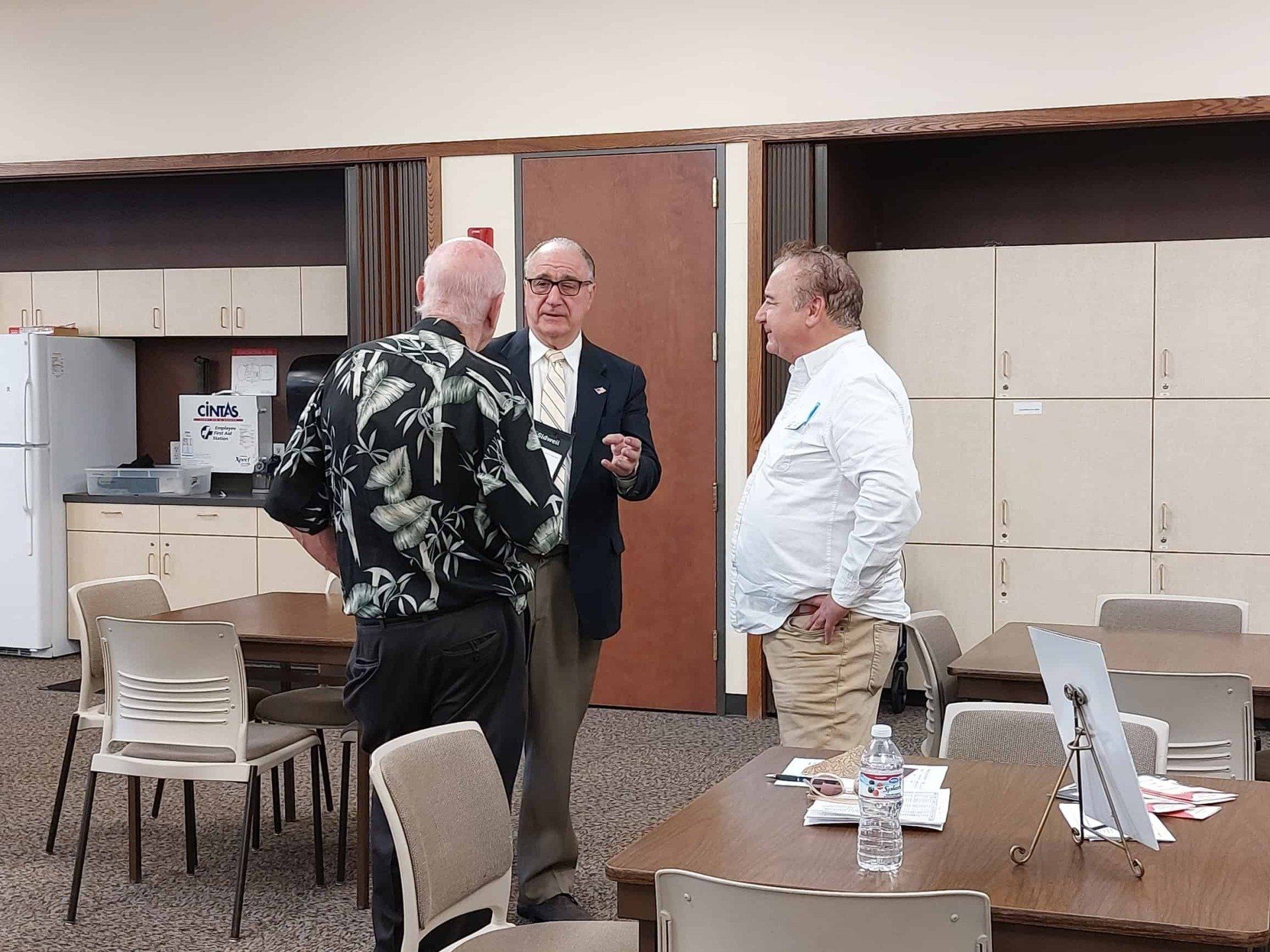 Three men talking in a room with tables, chairs, and folded wall partitions.