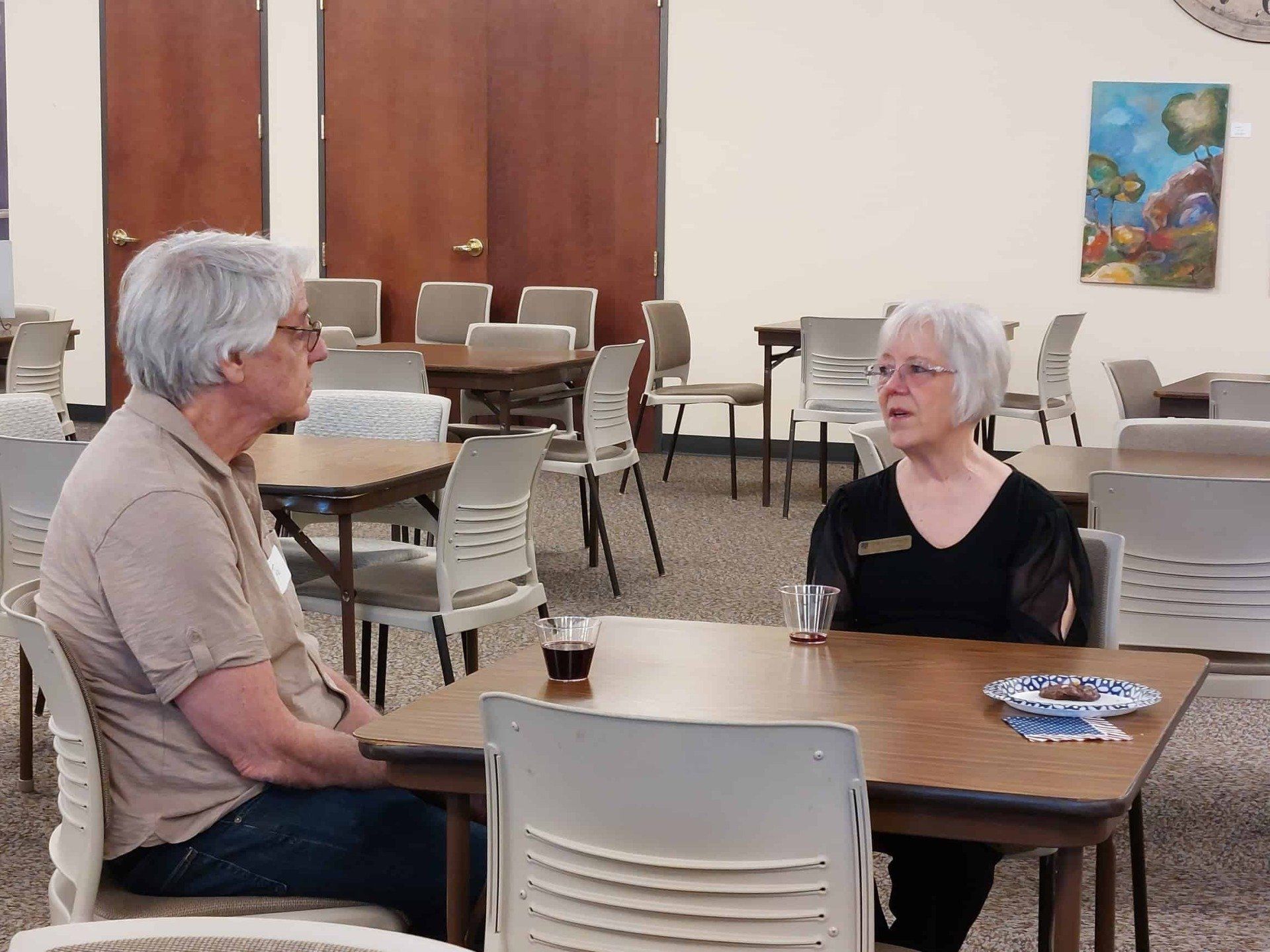 Two people talking at a round table in a large meeting room with chairs and wooden doors.