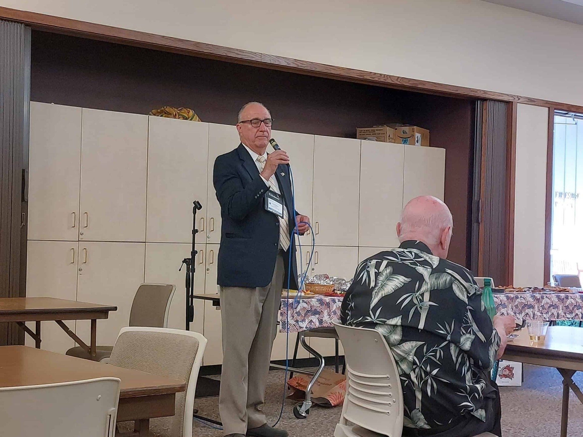 Man speaking into a microphone in a community hall while a seated audience listens