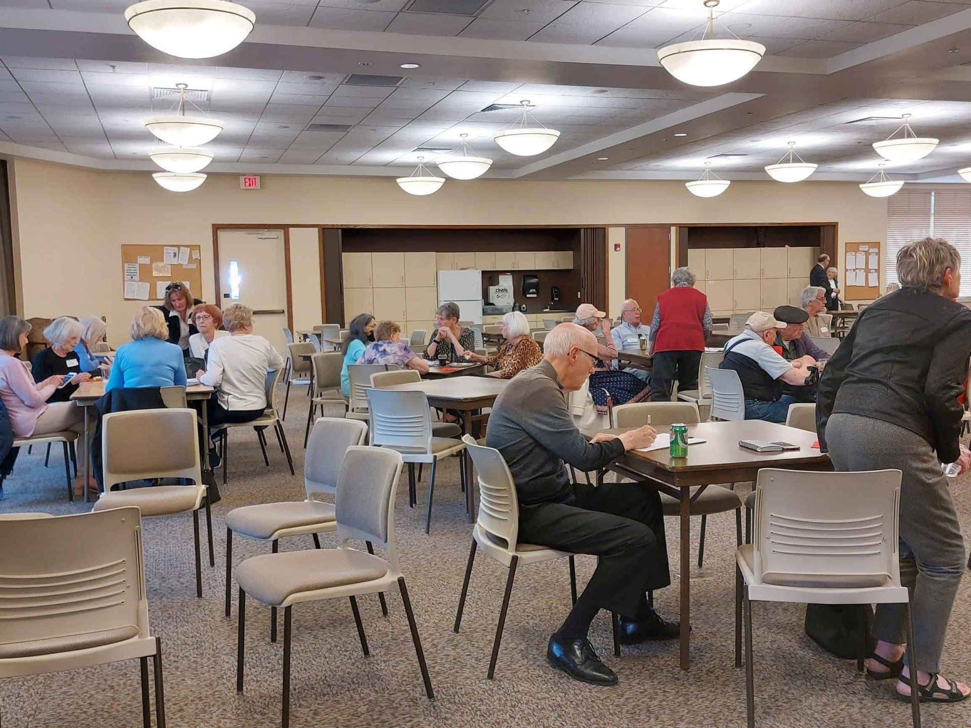Community hall with seated people at tables during a social gathering, under bright ceiling lights.