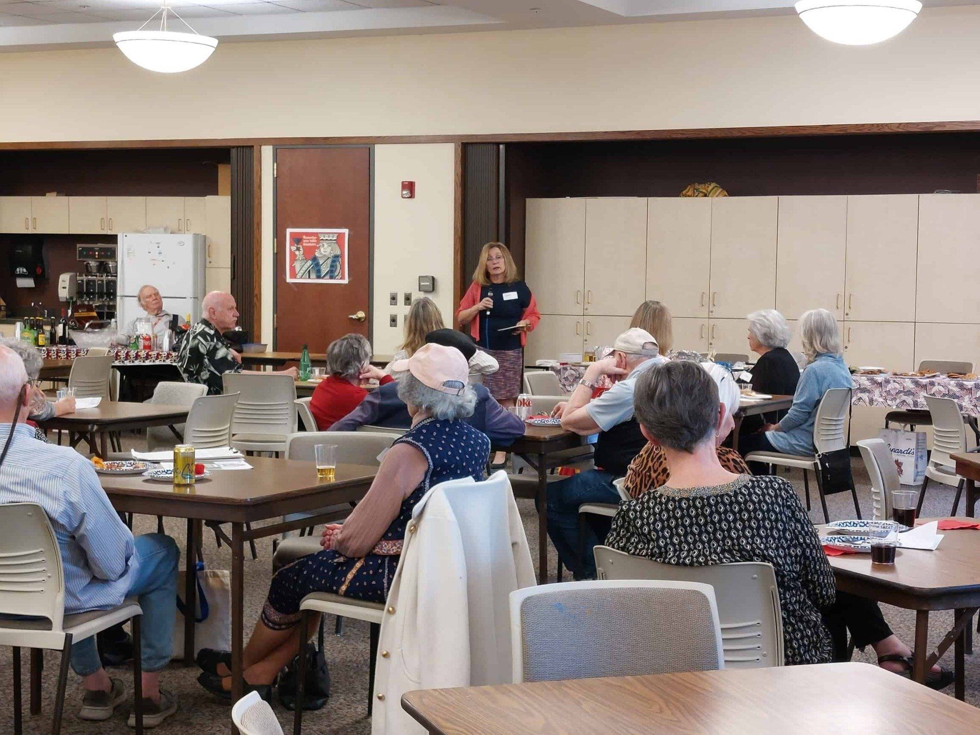 Community meeting in a dining hall, with people seated at tables and a speaker near the front.
