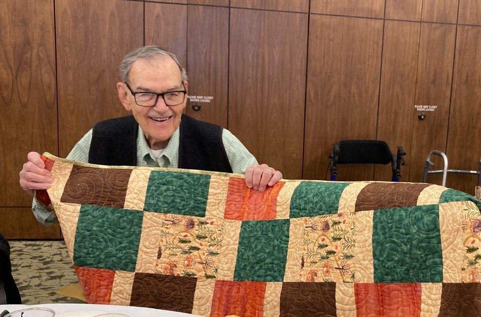 Smiling man holds up a colorful patchwork quilt in a wood-paneled room.