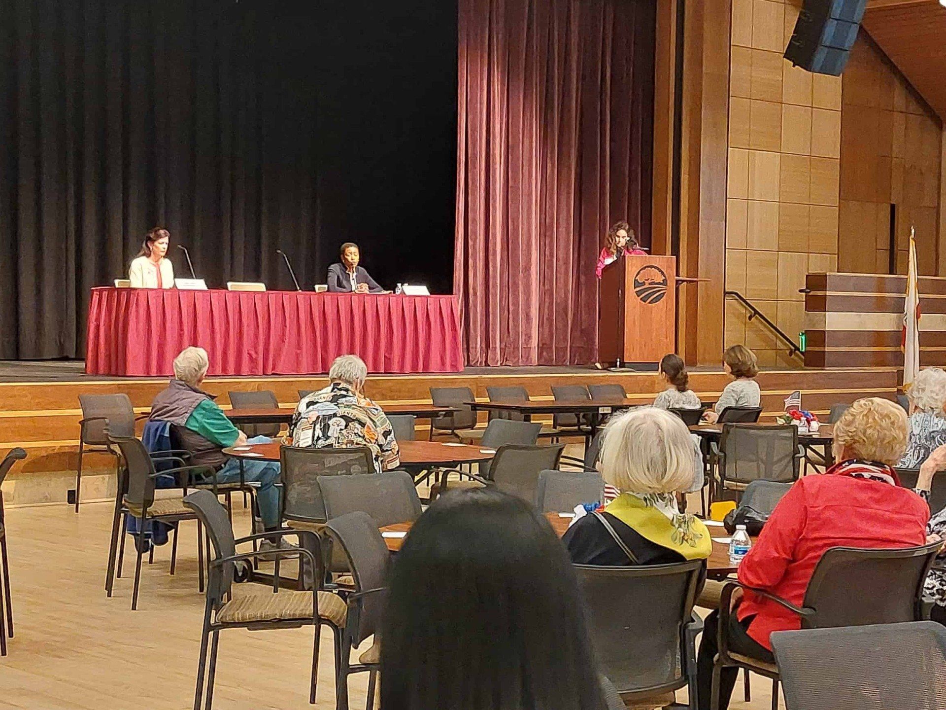 Audience seated in a hall facing a panel on a stage with red tablecloths and a podium