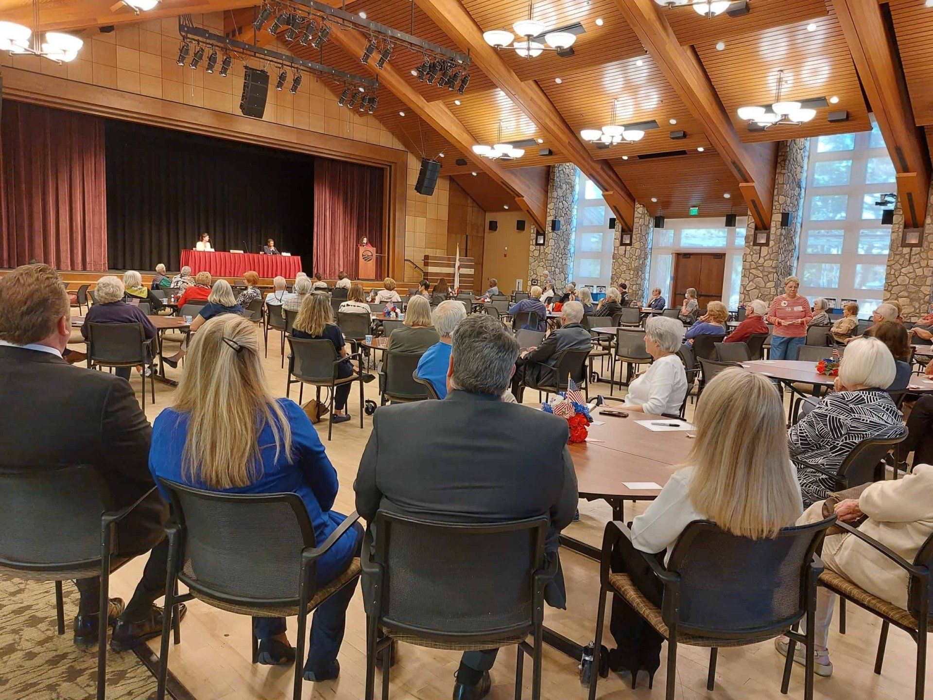 Audience seated in a wood-paneled hall facing a stage with red-draped tables and windows at the back