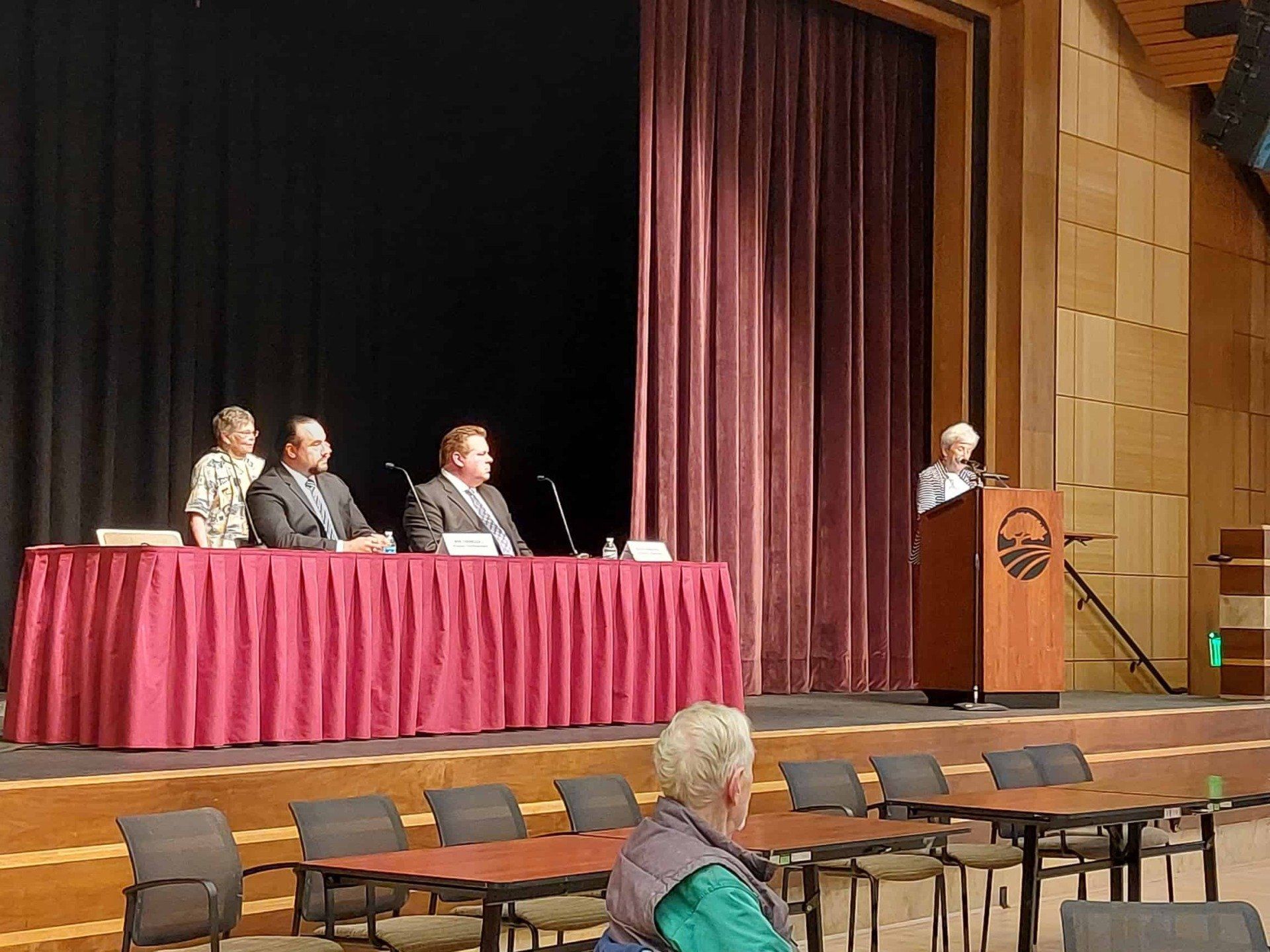 Panel discussion on stage with seated speakers and a presenter at a podium in a theater