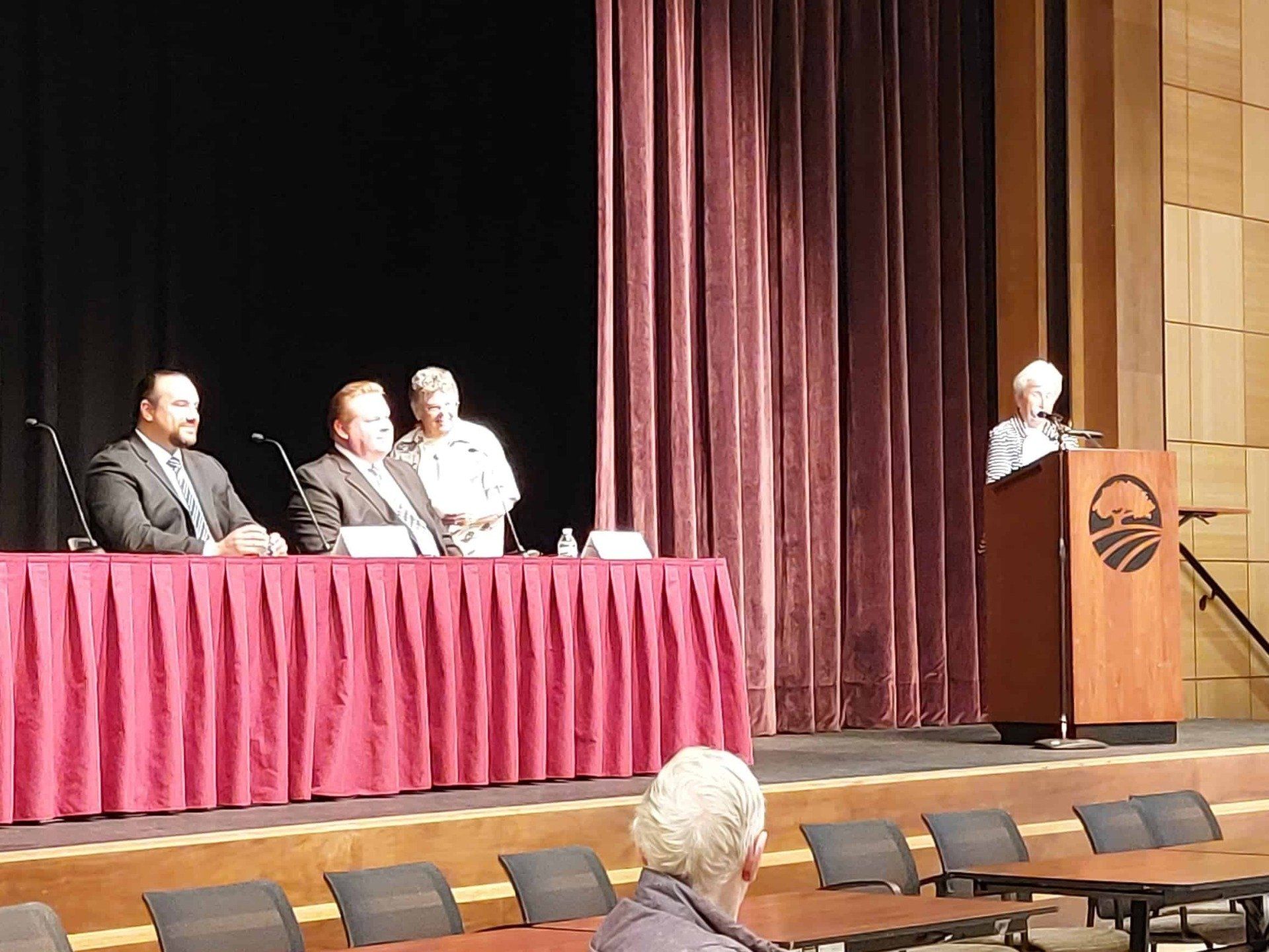 Panel discussion on stage with four speakers seated at a maroon-draped table and one at a podium in an auditorium