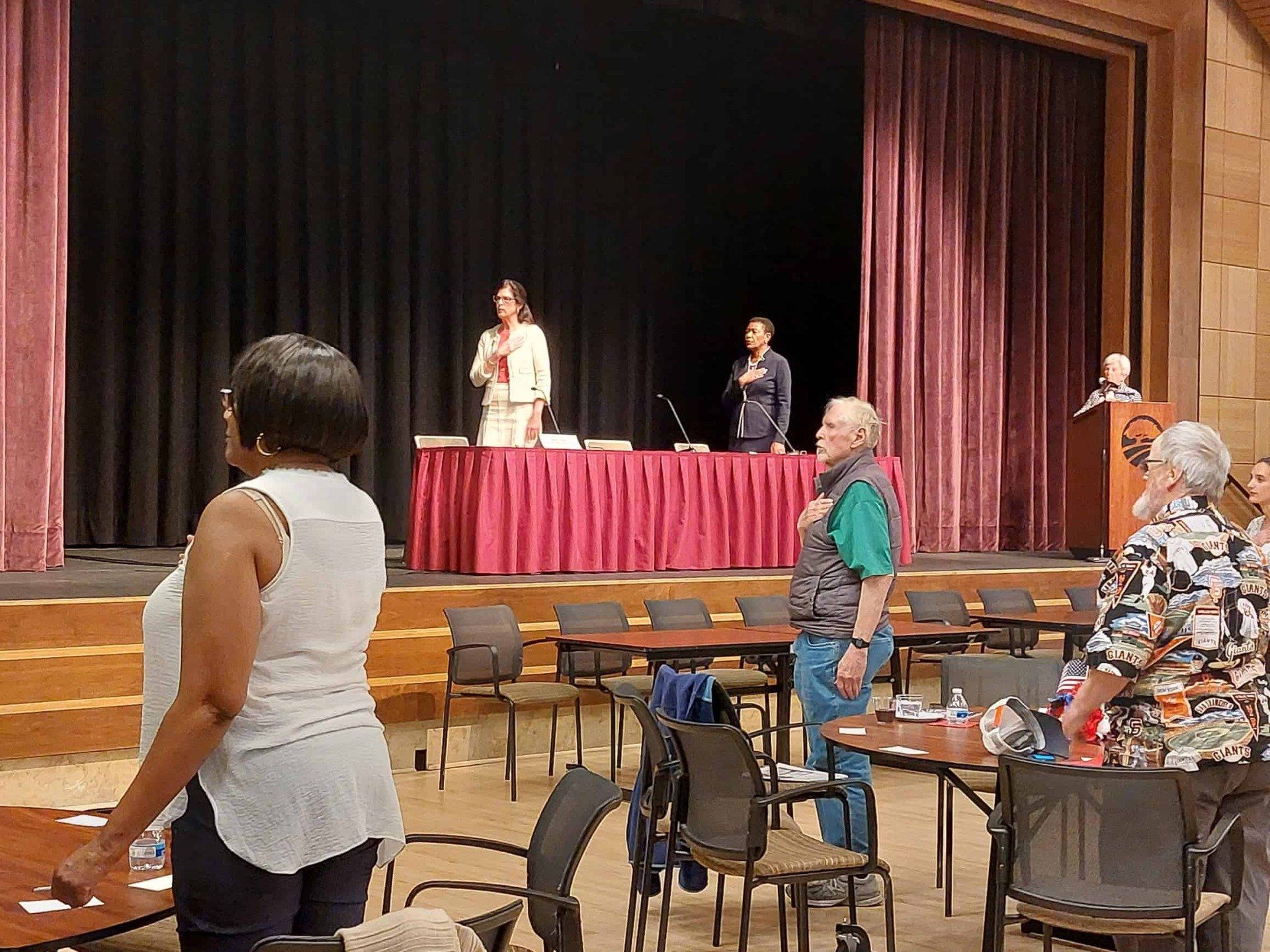 People seated in a hall facing a stage where two speakers stand at a podium and microphone.