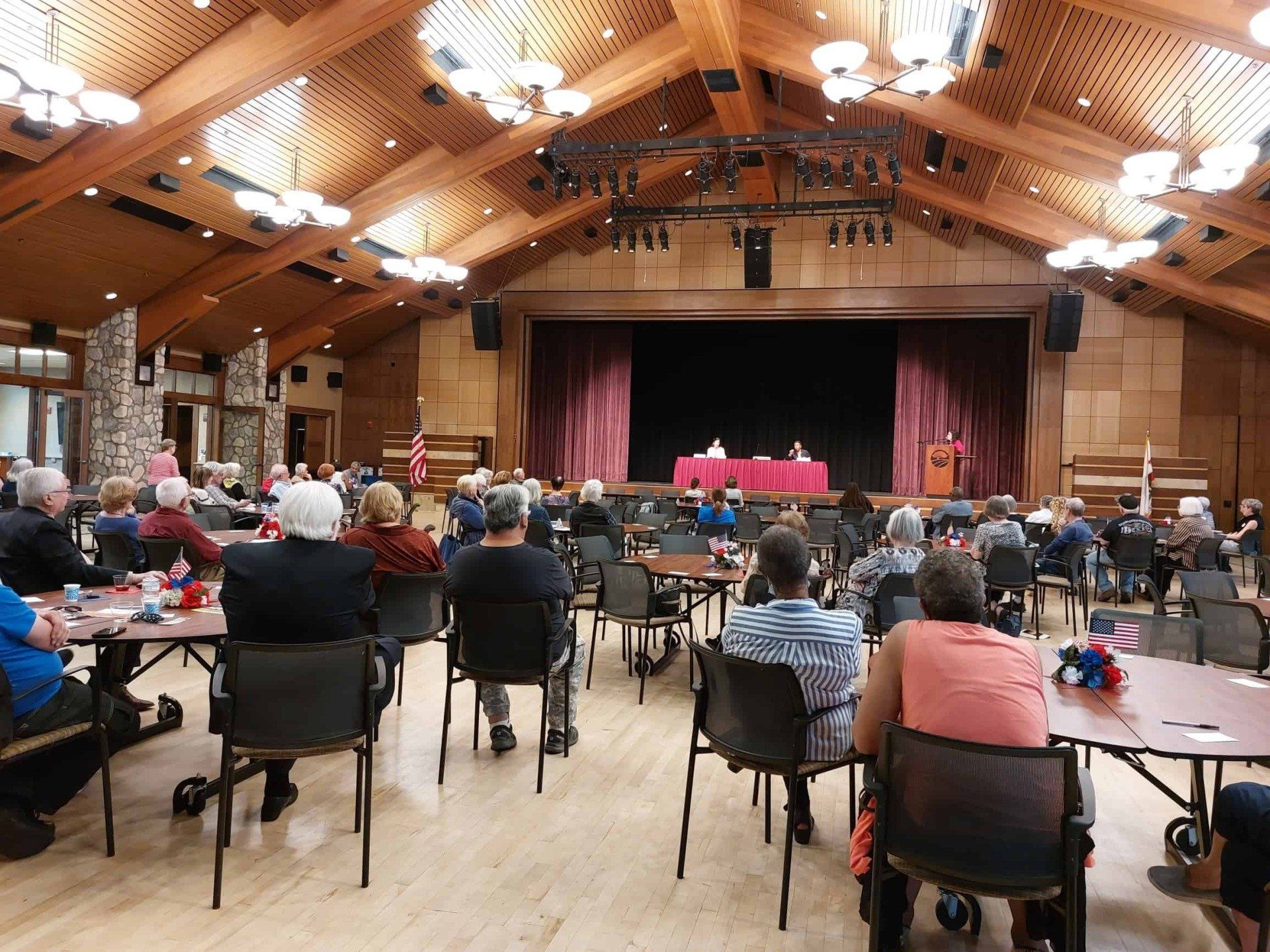 Audience seated in a wooden hall watching a performance on a small stage with red curtains