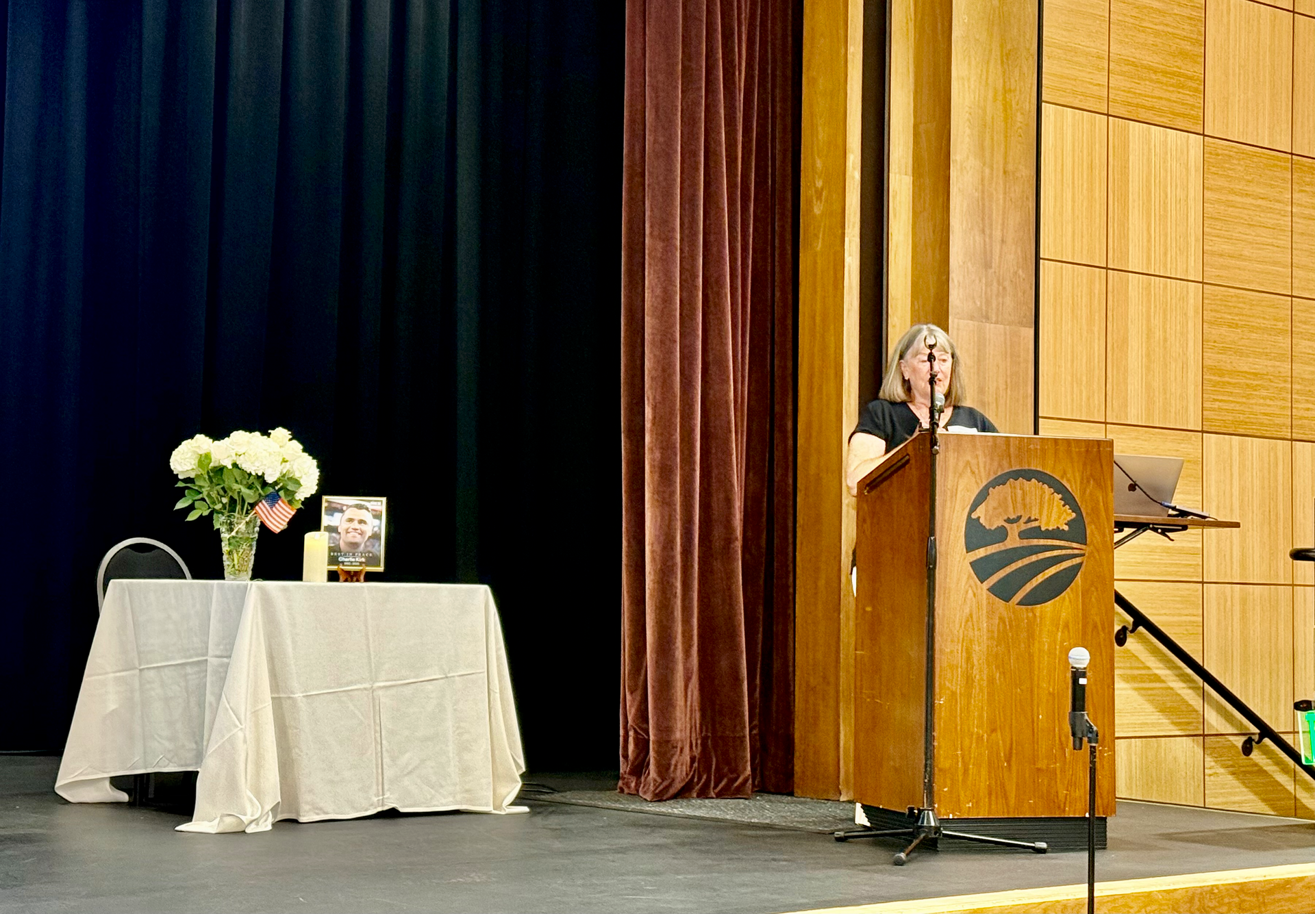 Speaker at podium on stage beside a small table with flowers and books, with draped curtains behind.