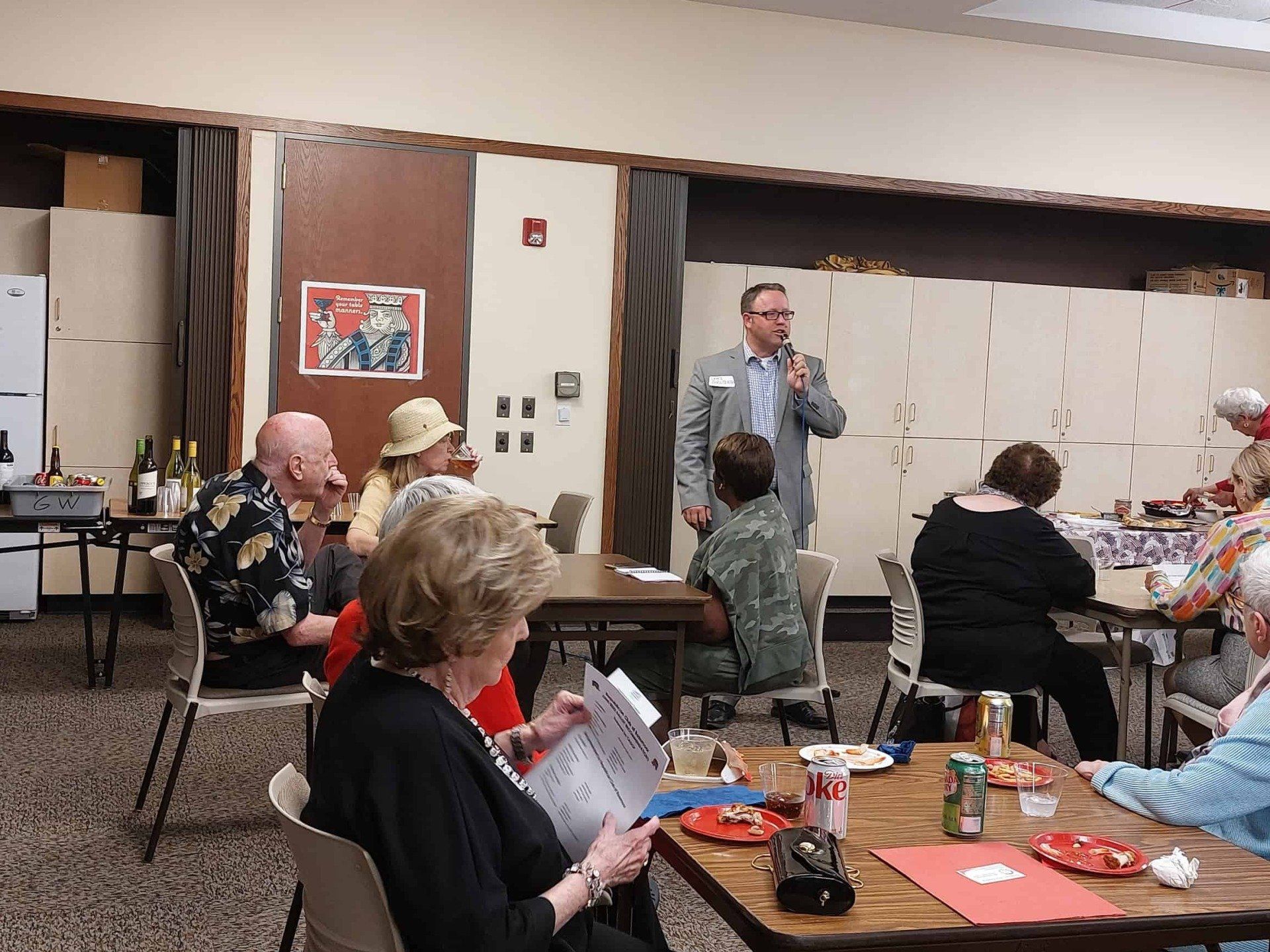 Community meeting in a hall, with a speaker at the front and seated attendees at tables.