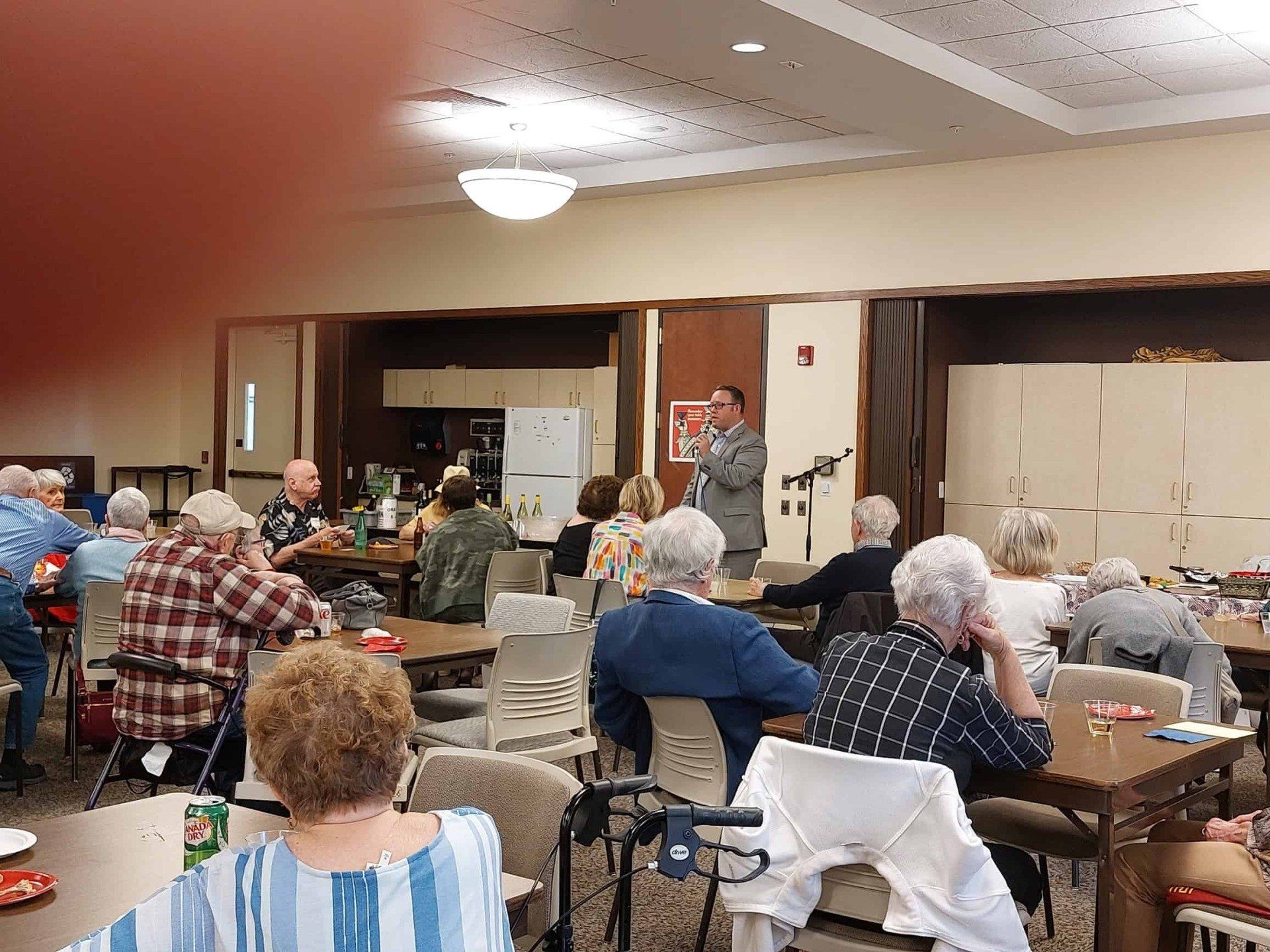 People seated at tables in a community hall, watching a speaker near the front.