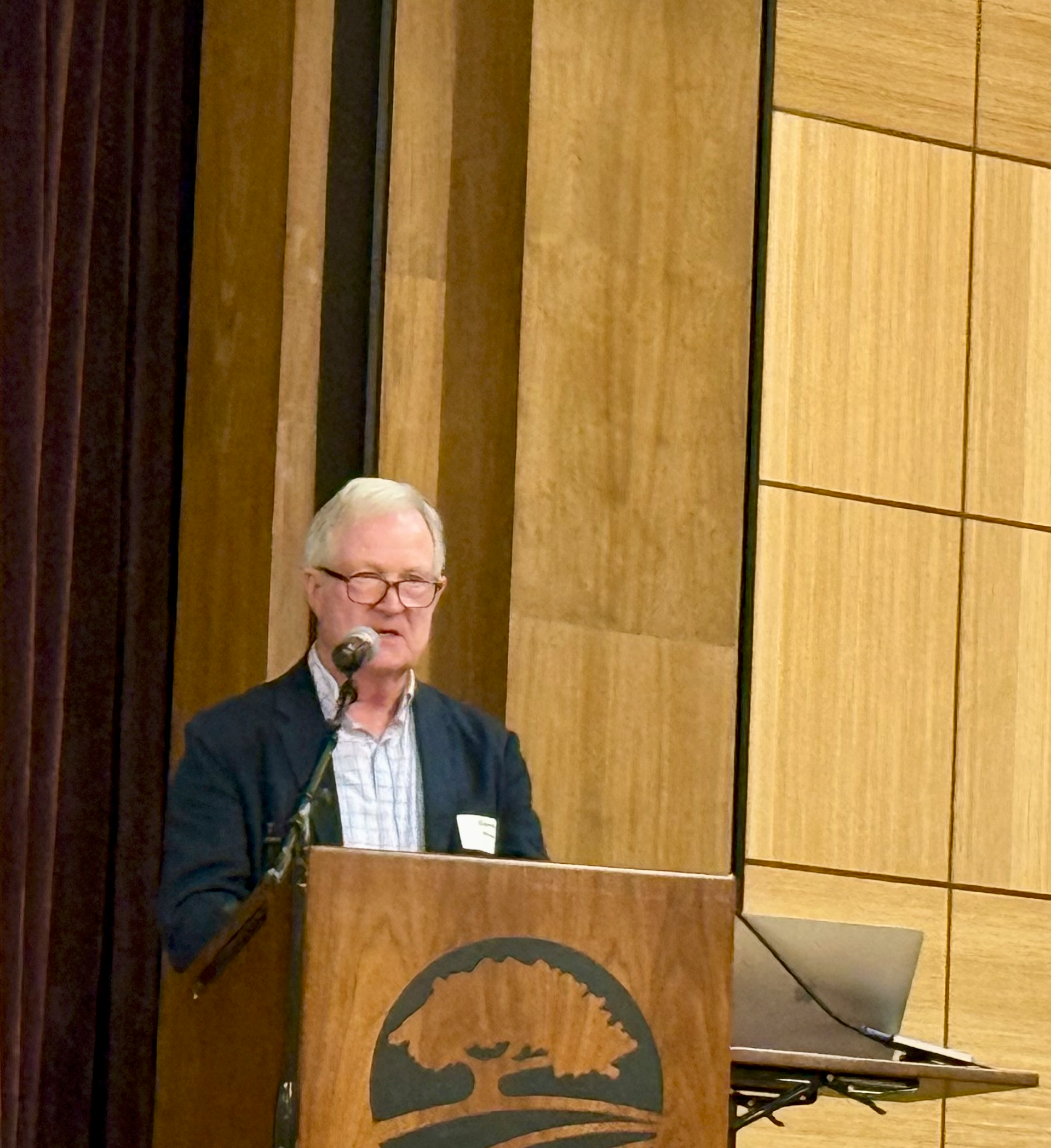 Speaker at a podium in a wood-paneled auditorium, speaking into a microphone with a laptop nearby.