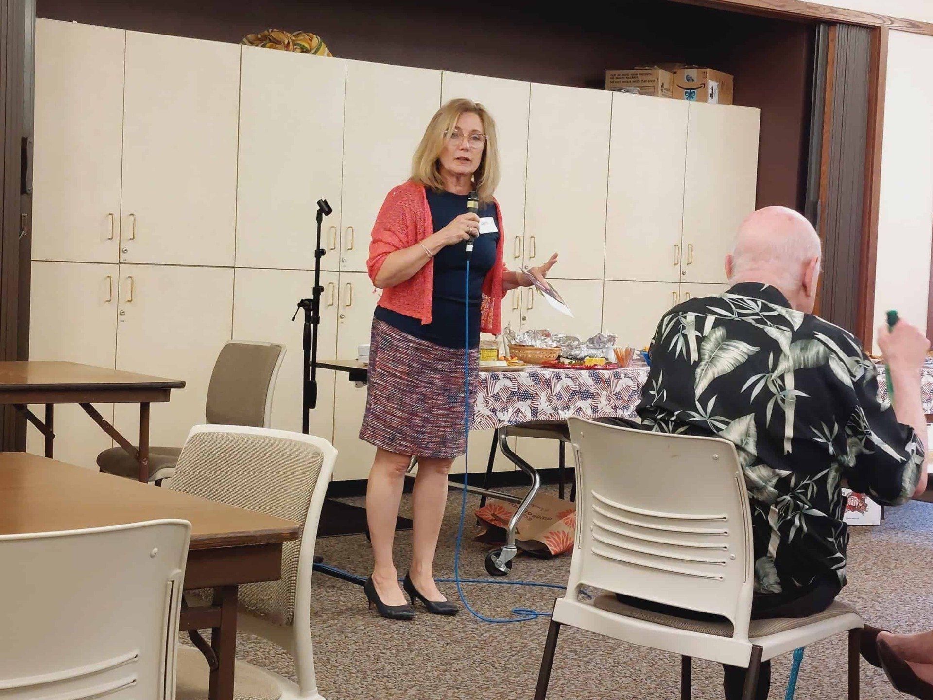 Woman speaking into a microphone at a meeting, with seated attendees and a table in a room