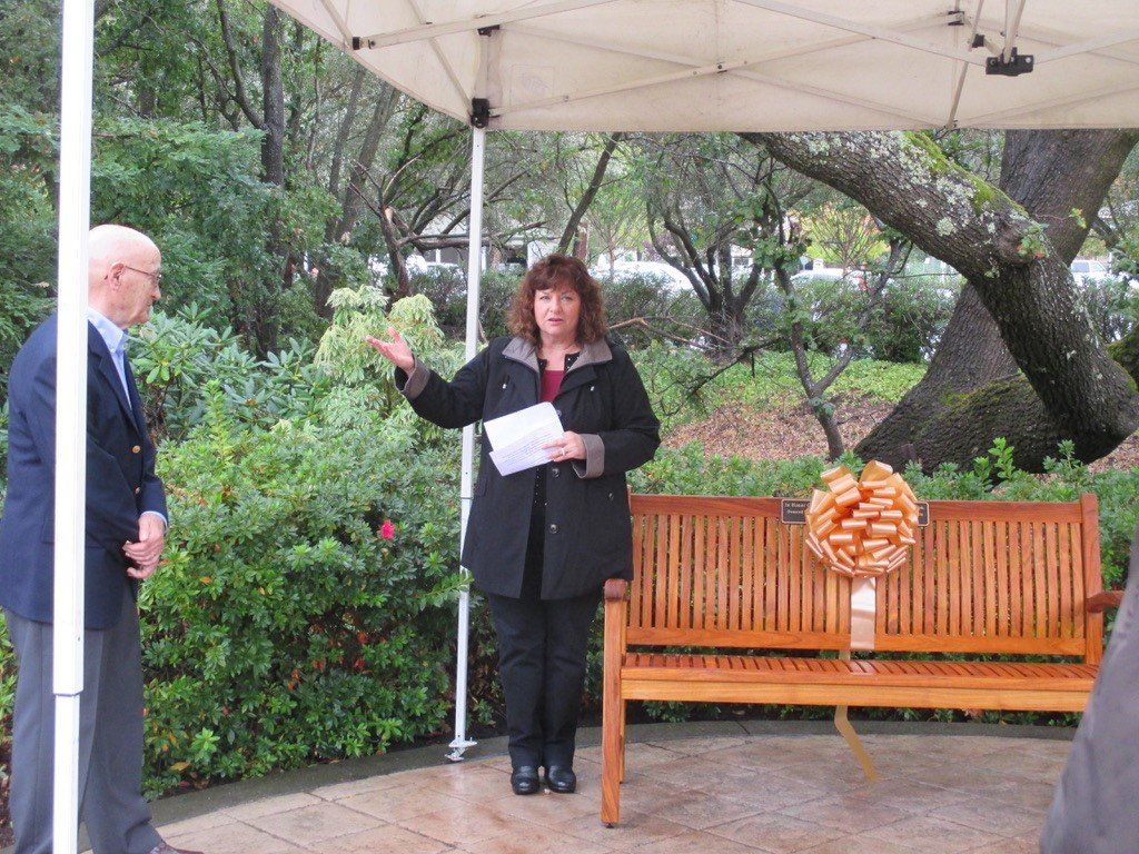 Woman speaking under a canopy beside a wooden bench in a garden, holding papers and gesturing toward a man.