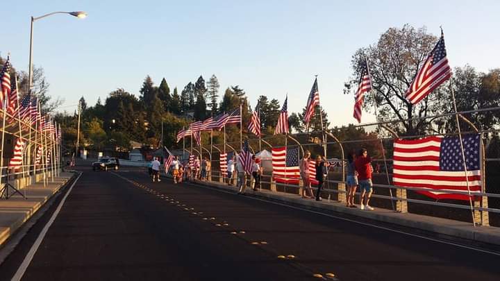 Bridge lined with American flags at sunset, with a few people standing along the roadside.