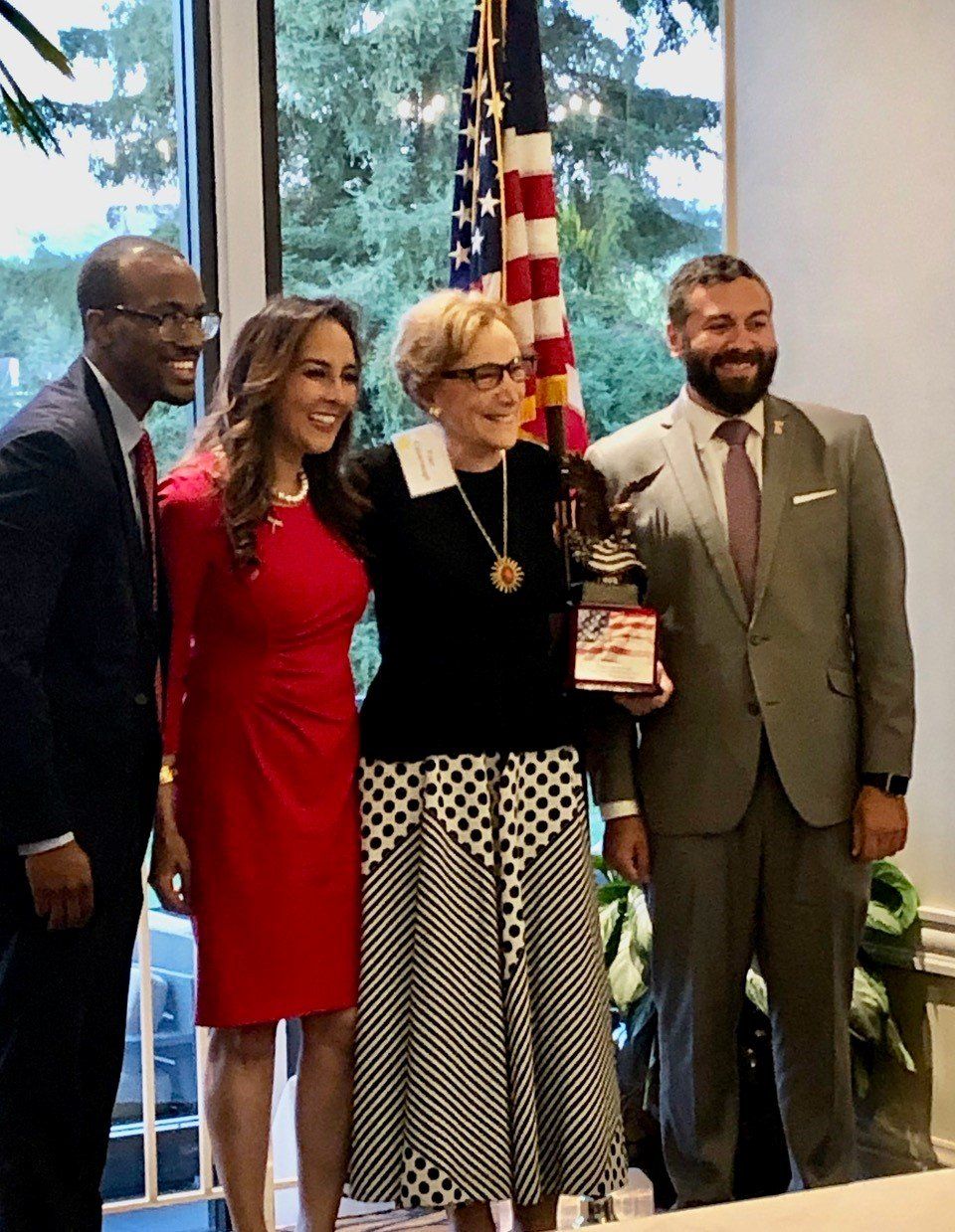 Four people posing indoors with an American flag; one holds an award plaque.