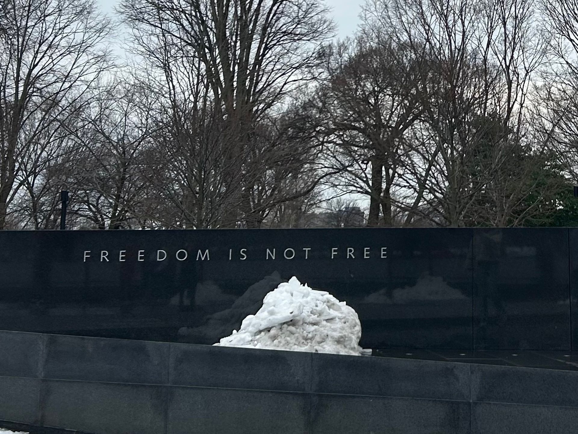 Snow piled against a black memorial wall inscribed “FREEDOM IS NOT FREE,” with bare trees behind it.