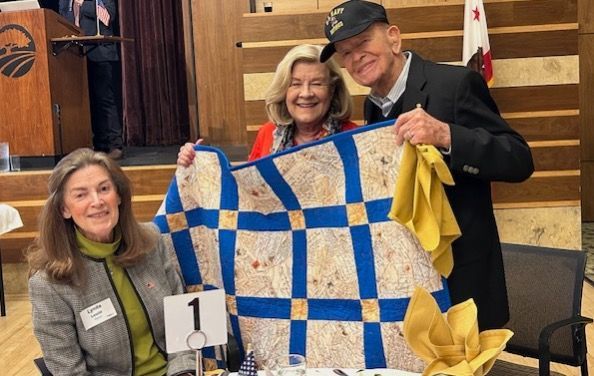 Three people pose indoors holding a blue-and-white quilt with yellow bow ties and a “1” tag.
