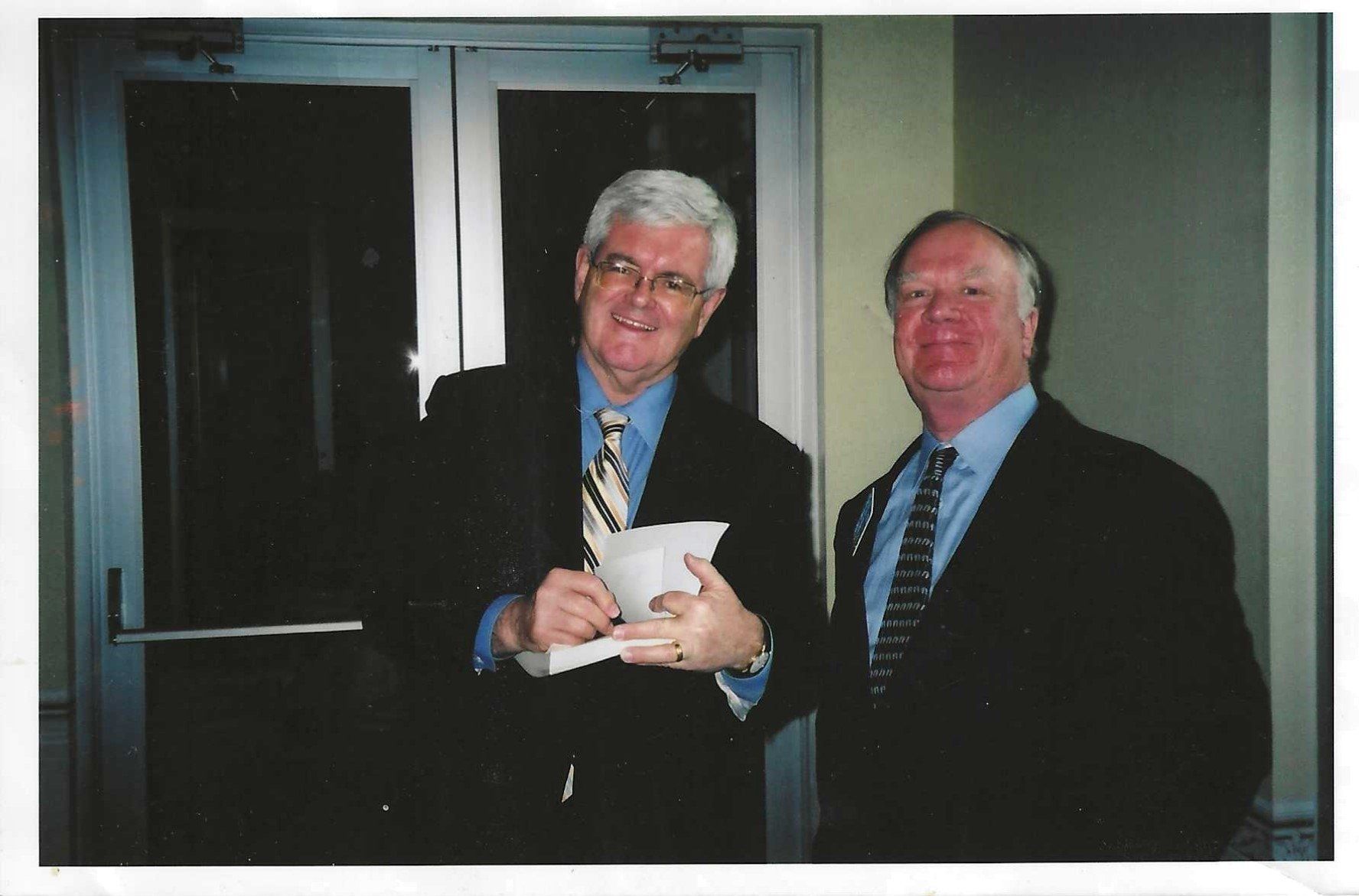 Two men in suits smiling indoors, holding a small object together
