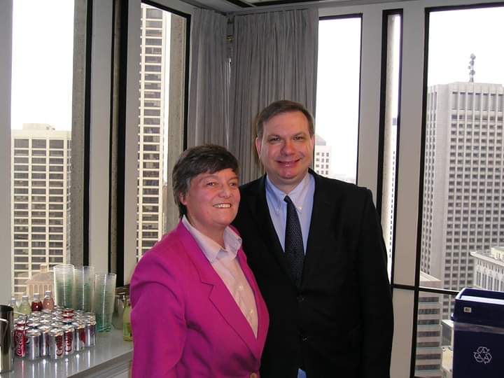 Two people in business attire smiling in an office with city buildings outside the windows