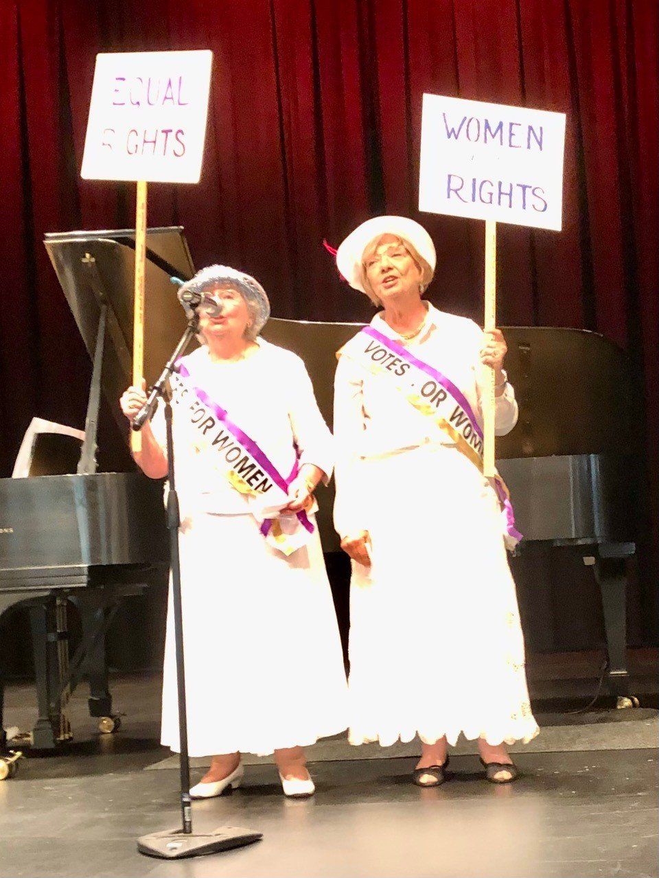 Two women in white dresses with sashes hold signs reading “Equal Rights” and “Women Rights” onstage.
