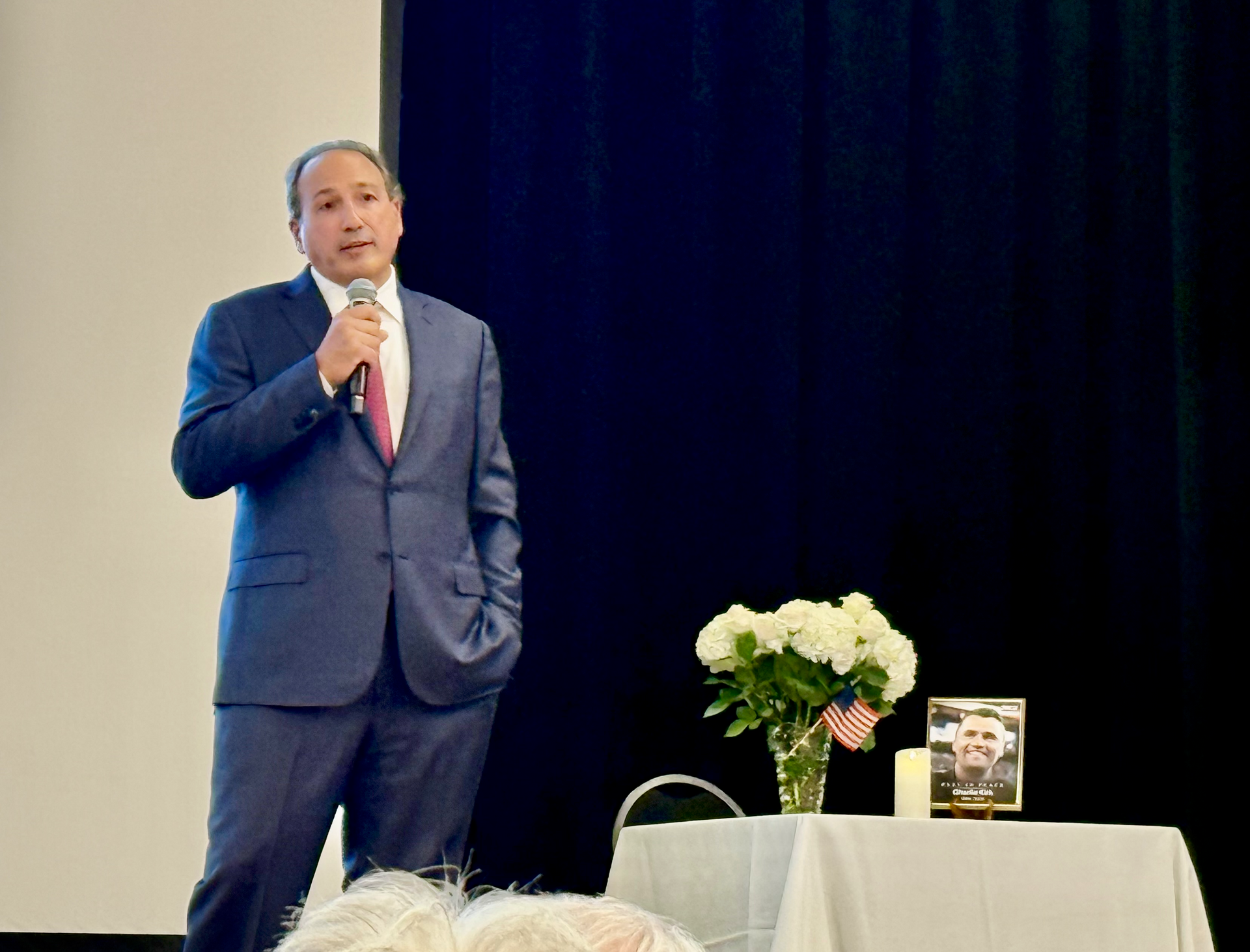Man in a blue suit speaking into a microphone beside a table with flowers and a book display onstage