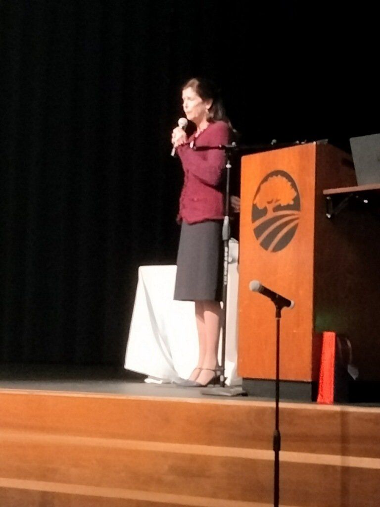 Woman speaking on a stage with a microphone beside a podium and microphone stand