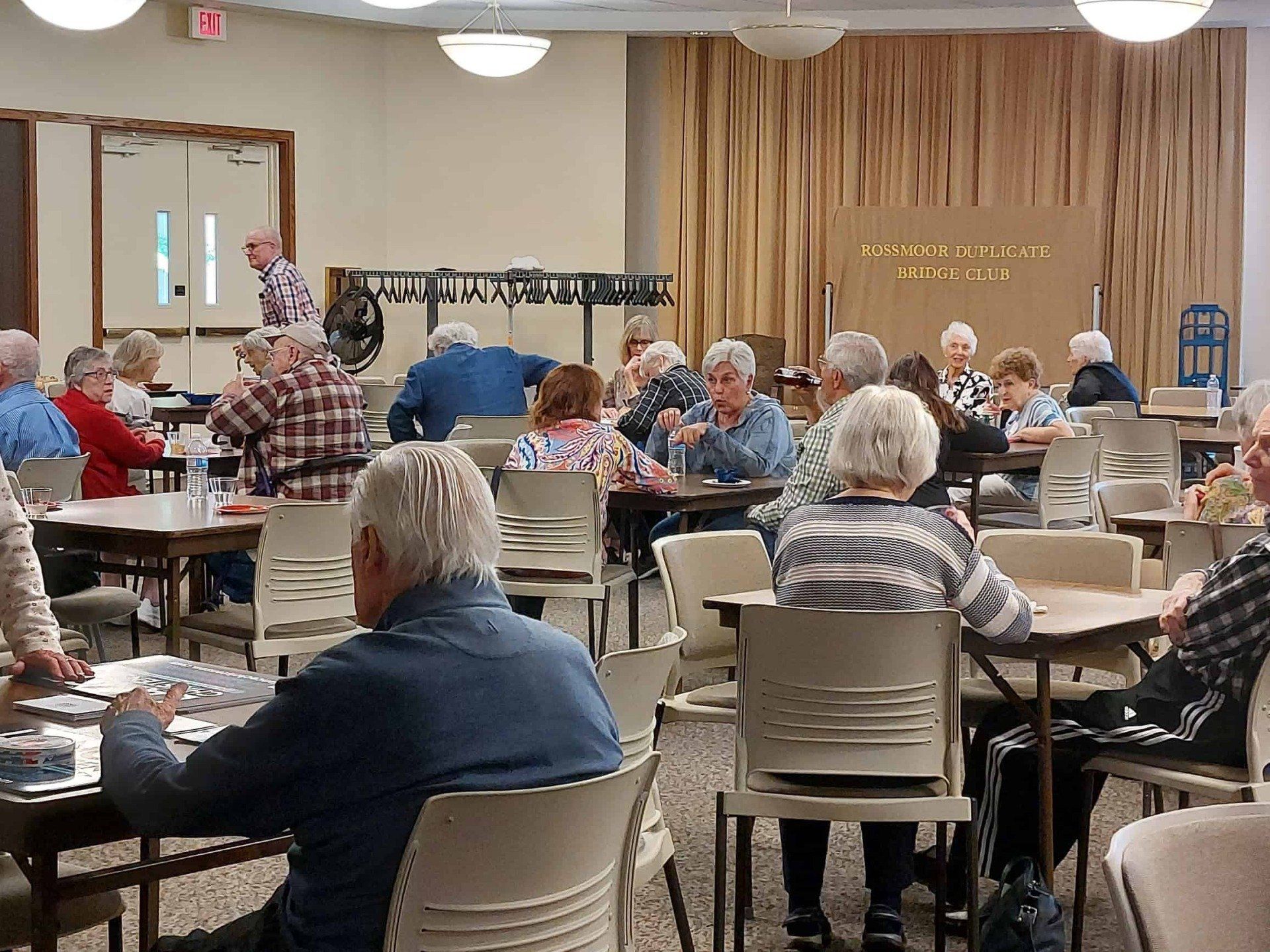 People seated at tables in a community hall, eating and chatting during a social gathering.