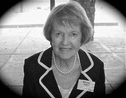 Smiling woman in a black jacket with white trim and pearl necklace, standing outdoors on a paved area.