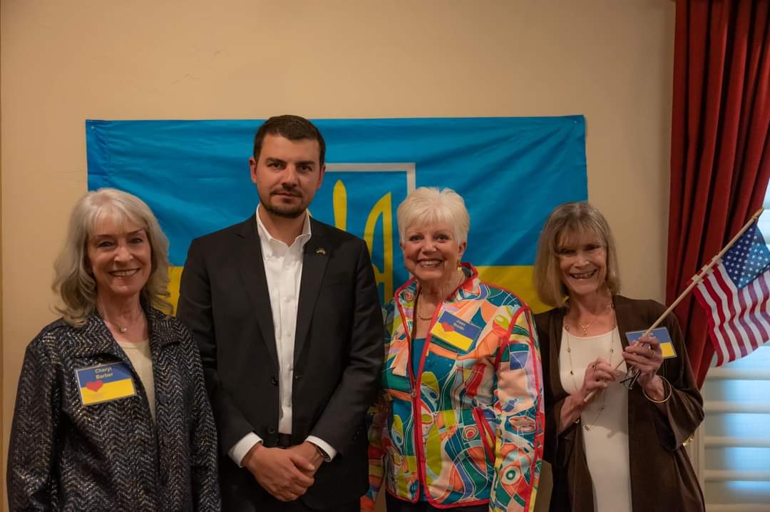 Four people posing indoors in front of a blue and yellow flag, one holding a small American flag.