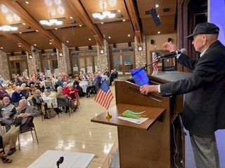 Speaker at podium addressing seated audience in a community hall with an American flag nearby