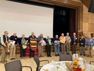 Group of people on stage holding gifts, standing in front of a large screen at an indoor event.