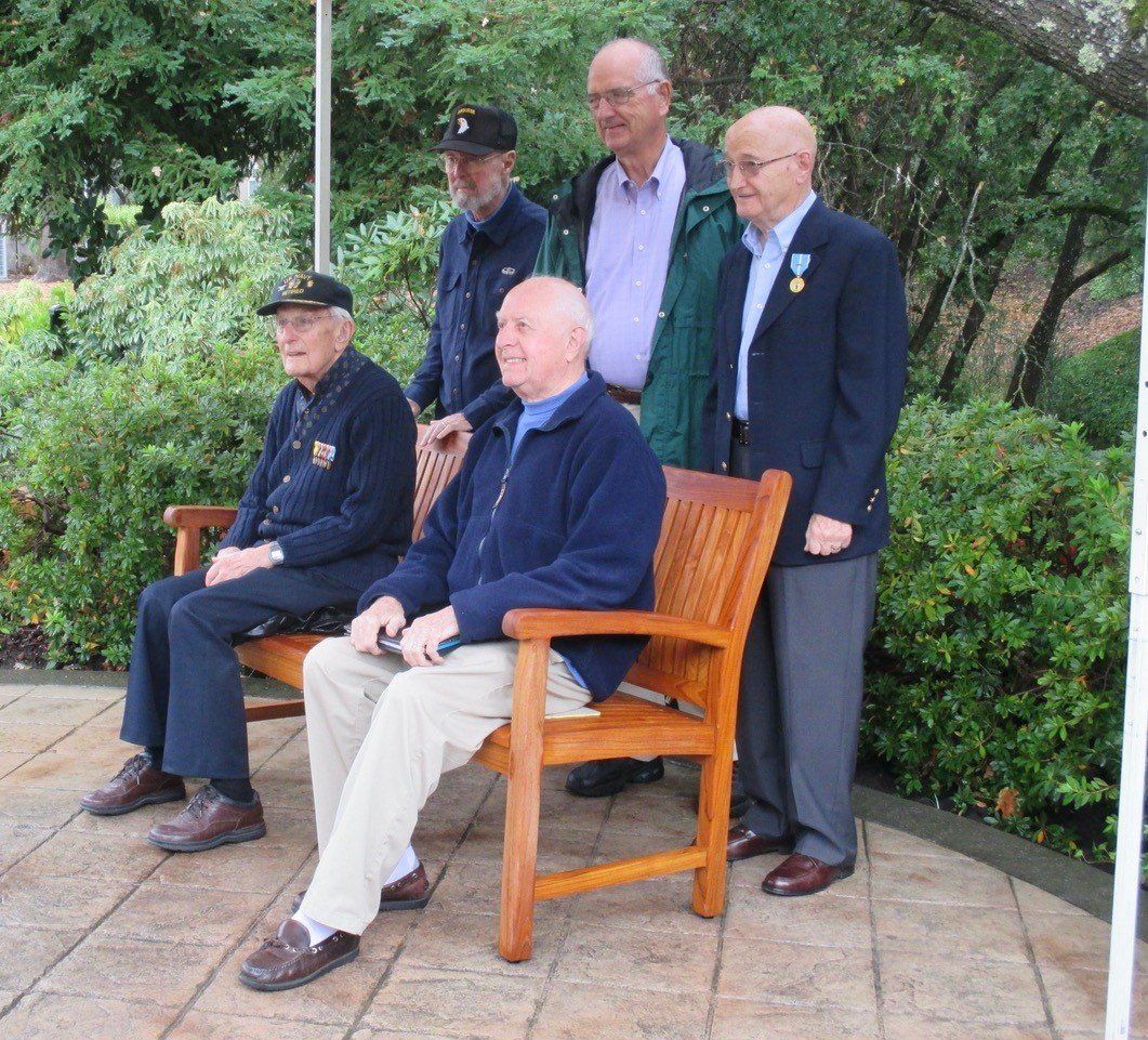 Five men in suits and caps pose on benches in a garden setting.