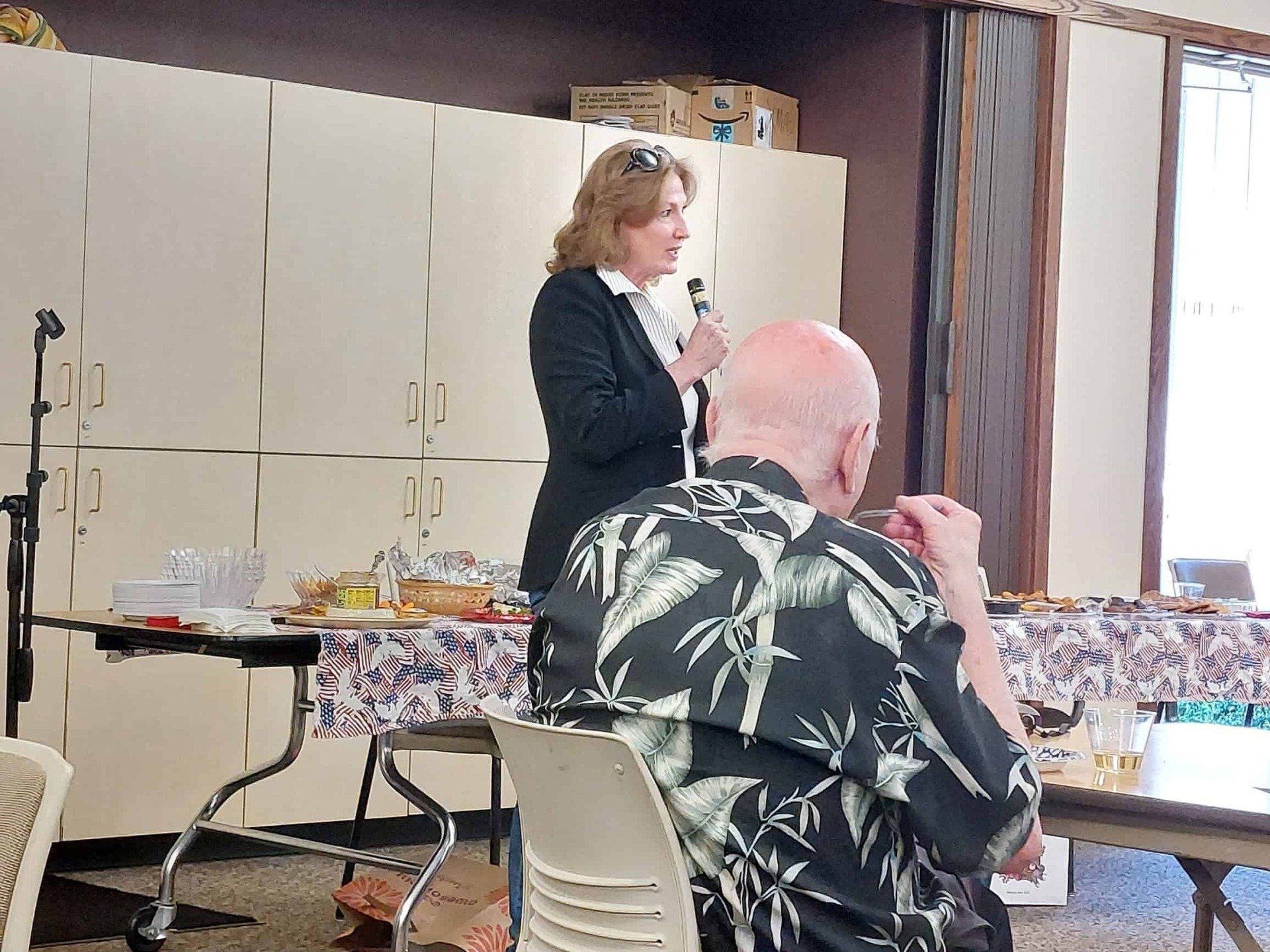 Woman speaks into a microphone at a decorated indoor gathering while a seated man listens in the foreground.