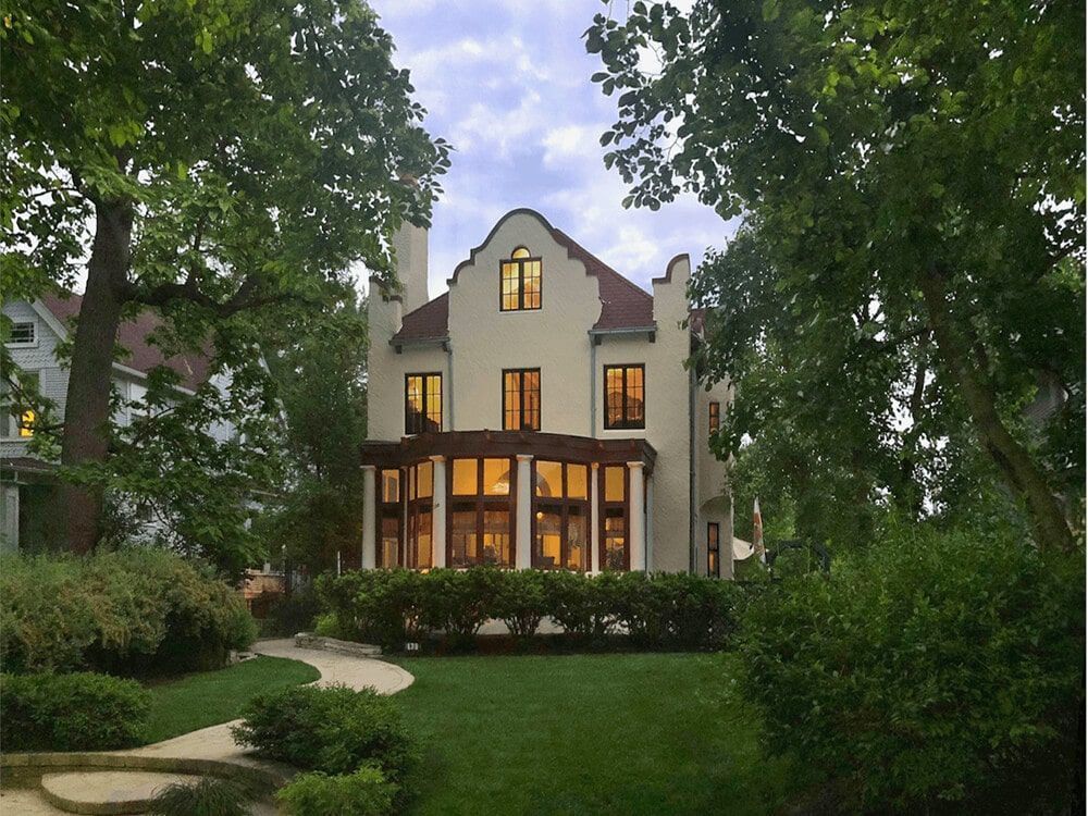 Elegant two-story house with warm lights, framed by trees and a lush green front yard