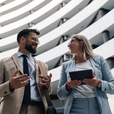 Two professionals in suits talking outside a modern curved building, one holding a tablet