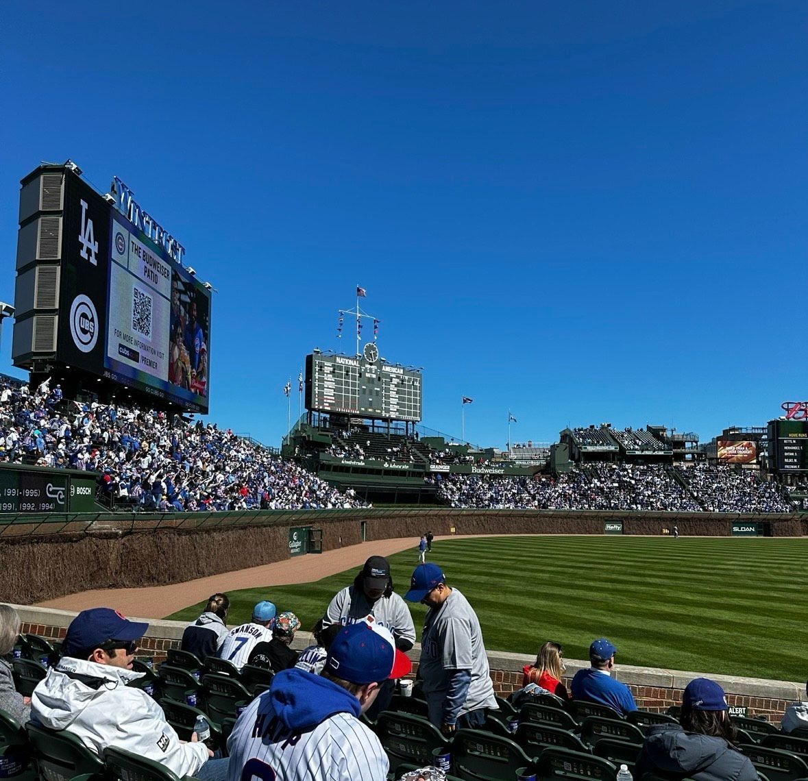 Baseball stadium with fans in winter coats under a bright blue sky, Chicago Cubs scoreboard visible