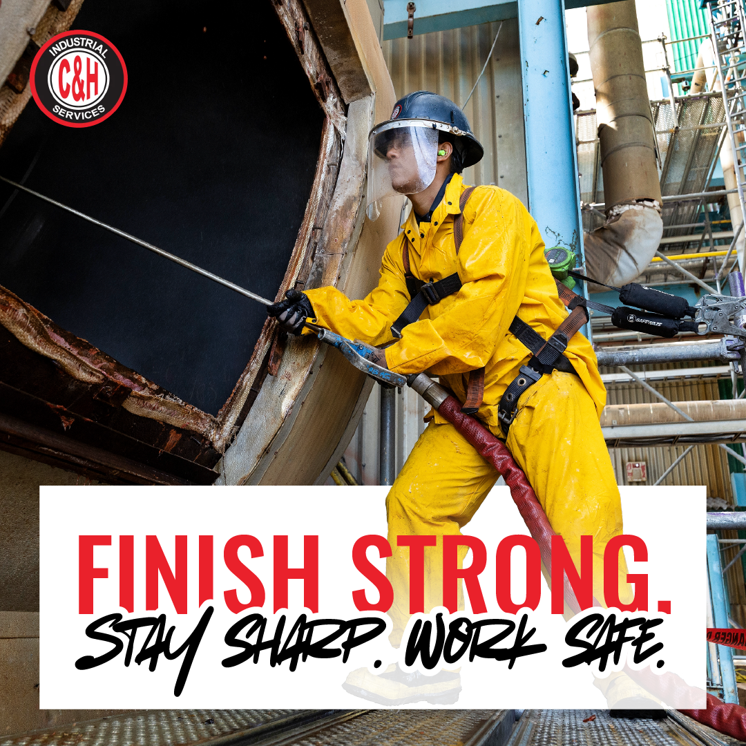 Worker in yellow safety gear using a hose at an industrial site with “FINISH STRONG. STAY SHARP. WORK SAFE.” poster