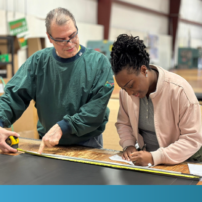 Two people measure and mark a long board on a worktable in a workshop