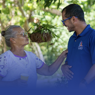 Two people smiling and talking outdoors, with one resting a hand on the other’s arm.