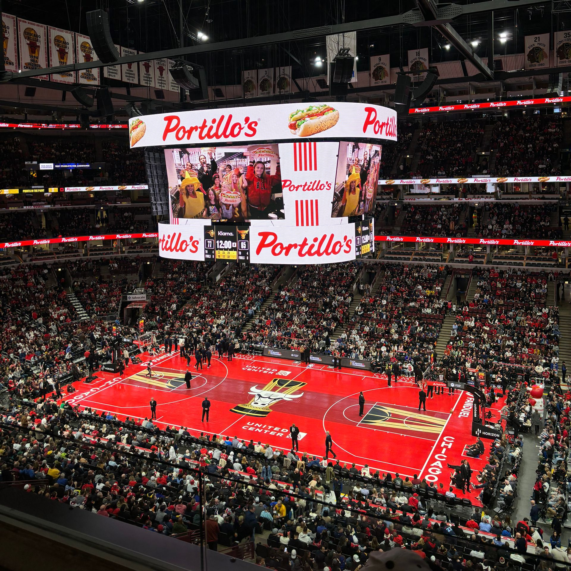 Basketball arena packed with fans, red court, and a large Portillo’s scoreboard display.