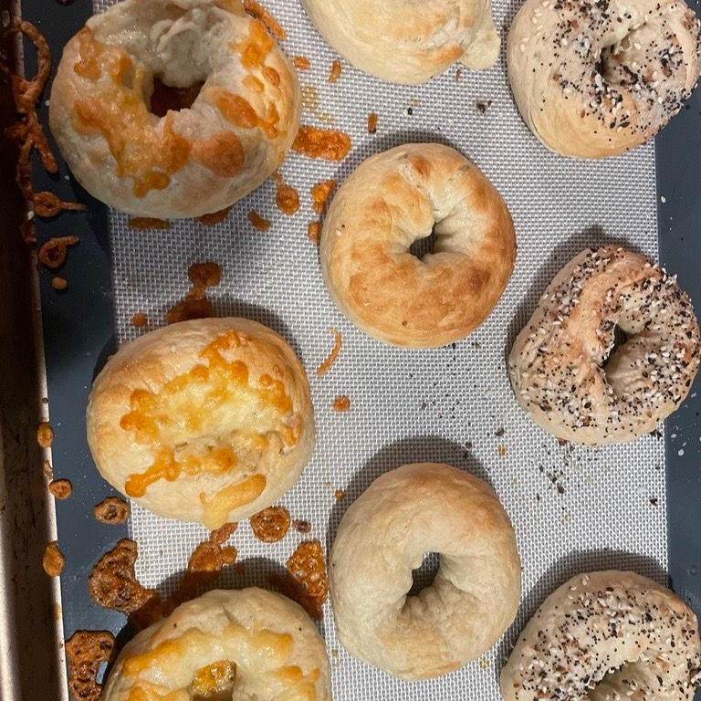 Assorted baked bagels on a tray, some topped with cheese, poppy seeds, and sesame seeds