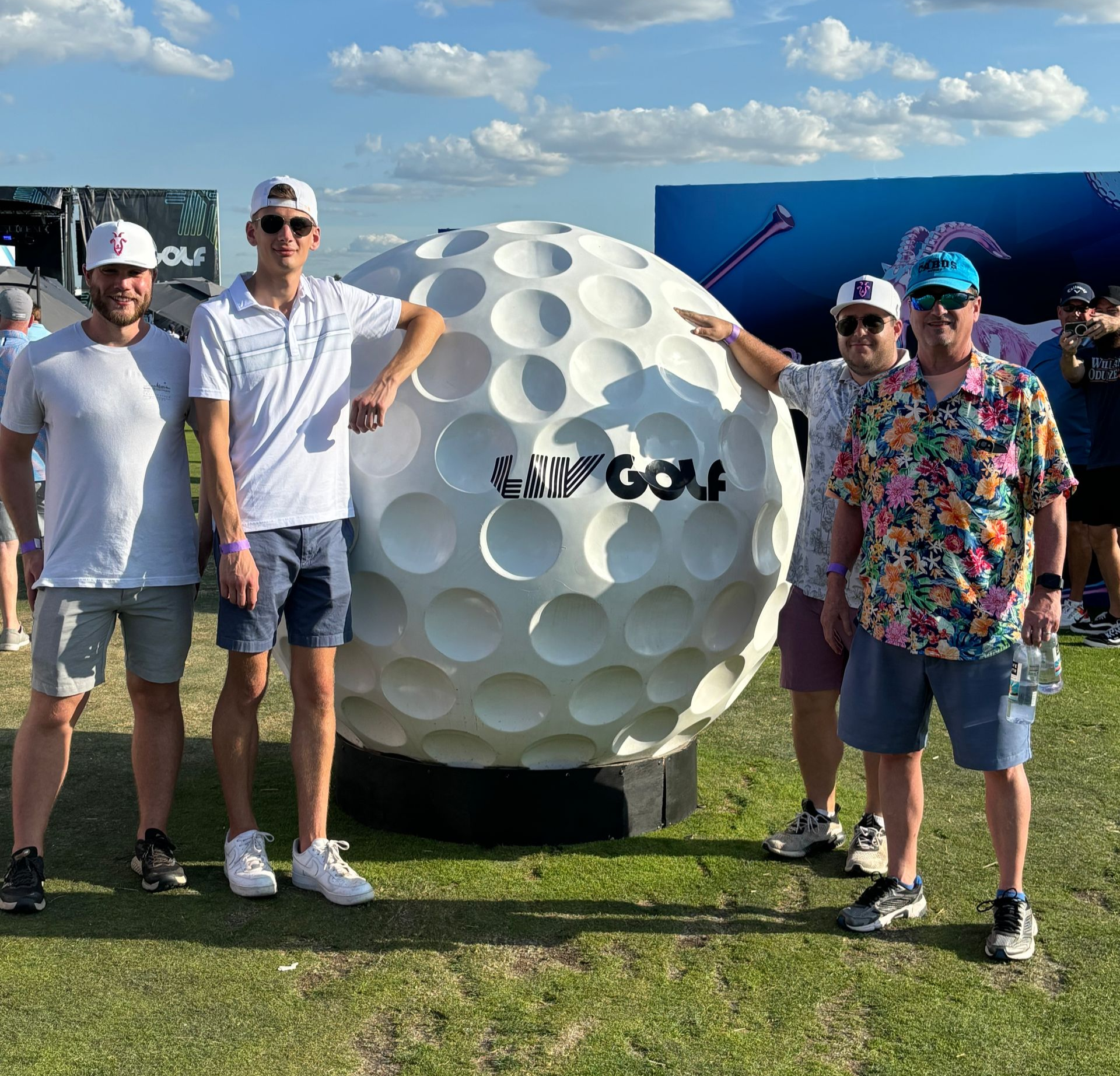 Four men posing beside a giant golf ball sculpture on a sunny grassy field.