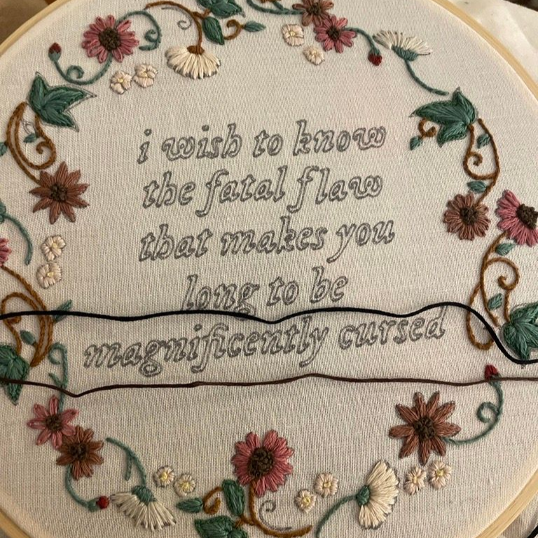 Embroidered hoop with floral wreath and quote: “I wish to know the fatal flaw that makes you long to be magnificently cursed”