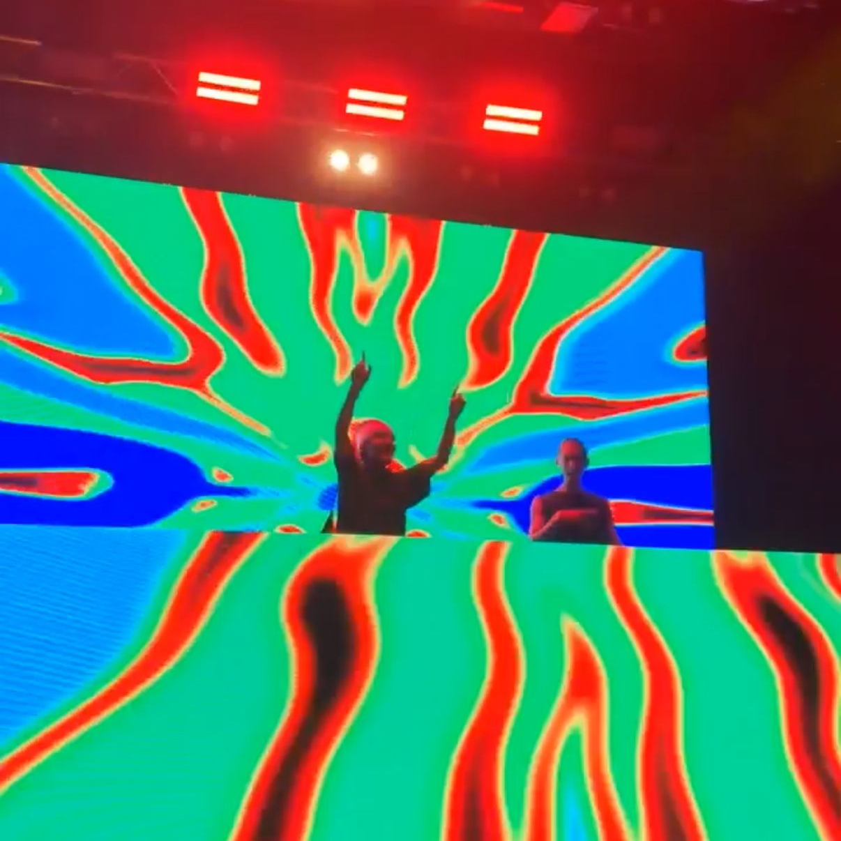 Two performers onstage before a large neon abstract LED backdrop with red lights overhead