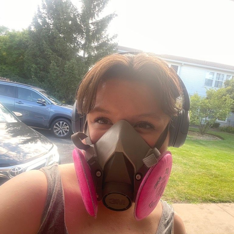 Selfie outdoors wearing a black-and-pink respirator mask, with a car and house in the background